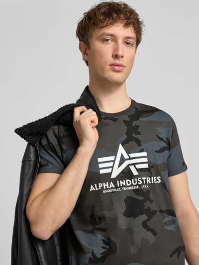 Männermodel trägt T-Shirt mit Label-Print von Alpha Industries, dunkle Camouflagefarben, Reine Baumwolle, Gerader-Schnitt, Rundhalsausschnitt, Rückenlänge bei Größe M 70 cm, Nahaufnahme Oberkörper.