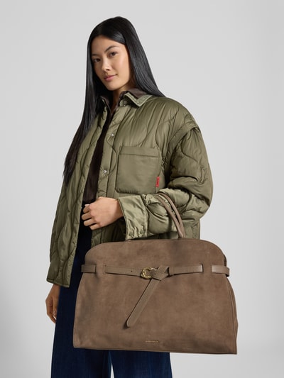 Frauenmodel trägt Handtasche mit Logo-Detail Modell 'Sabine' von Coccinelle in Braun, sichtbar genarbte Textur, Ganzkörperansicht von vorne. Geteiltes Hauptfach, Magnetverschluss, verstellbarer Schulterriemen, Innenfach mit Reißverschluss, Maße: 44x32x16 cm, Schulterriemen 106 cm.