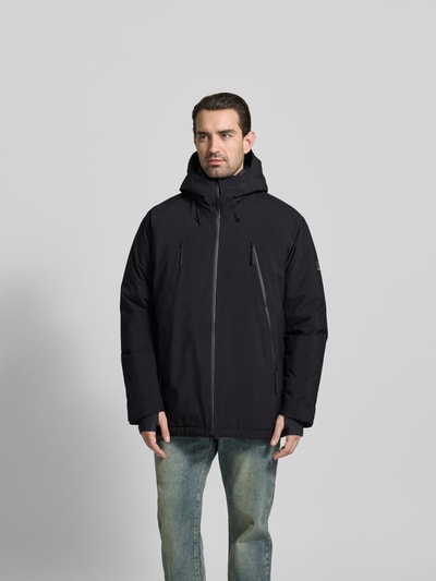Männermodel trägt Daunenjacke mit Daunen-Feder-Füllung von MM6 Maison Margiela in Schwarz, locker, Kapuze, Label-Print, Ganzkörperansicht von vorne. Rückenlänge 87 cm, Schulterbreite 57 cm, Ärmellänge 38 cm, Zweiwege-Reißverschluss, Reißverschlusstaschen, verstellbare Ärmelriegel, Tunnelzug im Bund.