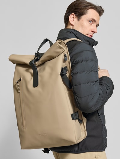 Männermodel trägt Rolltop Rucksack mit Ritszak van Rains in beige mit zwarte details, karabijnhaak, draagband en verstelbare bandjes, Ganzkörperansicht Rückansicht. Breedte 31 cm, lengte 61 cm, diepte 15 cm, schouderbandlengte 74 cm.