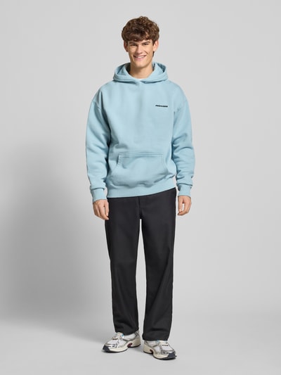Męski model nosi Oversized Hoodie mit Logo und Känguru-Tasche von Pegador, bawełniany miks, oversize, logo, kieszeń kangurka, długość tyłu w rozmiarze M: 75 cm, długość rękawa w rozmiarze M: 74 cm, jasnoniebieski, czarne spodnie, białe buty, gładka faktura, całkowity widok z przodu.