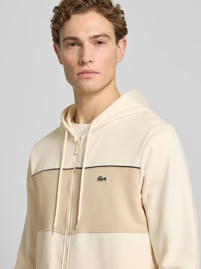 Männermodel trägt Regular Fit Sweatjacke aus reiner Baumwolle von Lacoste in Beige mit Kontraststreifen, Logo und Kapuze, Nahaufnahme Oberkörper.