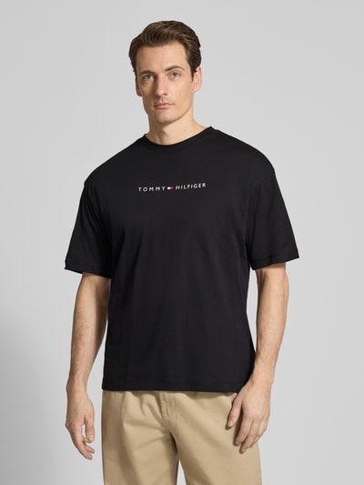 Männermodel trägt Regular Fit T-Shirt aus reiner Baumwolle von Tommy Hilfiger, unifarben in Schwarz, kurzer Arm, Label Print, Ganzkörperansicht von vorne.