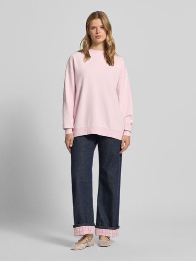 Frauenmodel trägt Oversized Sweatshirt mit rückseitigem Statement-Print von OH APRIL in Hellrosa, kombiniert mit weiter Jeans, Ganzkörperansicht von vorne. Reine Baumwolle, gerippter Rundhalsausschnitt, überschnittene Schultern, Logo-Stitching, Modell 'Tomato Girl', lässiger Oversized Fit.