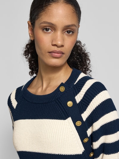 Frauenmodel trägt Regular Fit Strickpullover aus reiner Baumwolle von Tommy Hilfiger, gestreift in Blau und Creme, Zierknöpfe, Allover-Muster, Logo-Applikation, Rundhalsausschnitt, Materialverarbeitung Strick, Nahaufnahme Oberkörper.