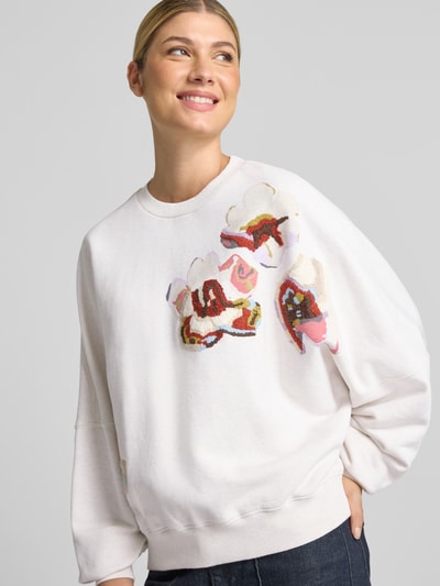 Vrouwenmodel draagt oversized sweatshirt uit puur katoen Model 'ASTERA' van DRYKORN, ribboorden, bloemmotief, witte en roze tinten, nahaufnahme bovenlichaam.