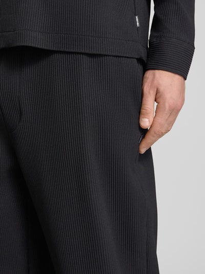 Männermodel trägt Hose mit elastischem Bund in Plissee-Optik von REVIEW, schwarzer Material-Mix mit Stretch-Anteil, Nahaufnahme Oberschenkel.