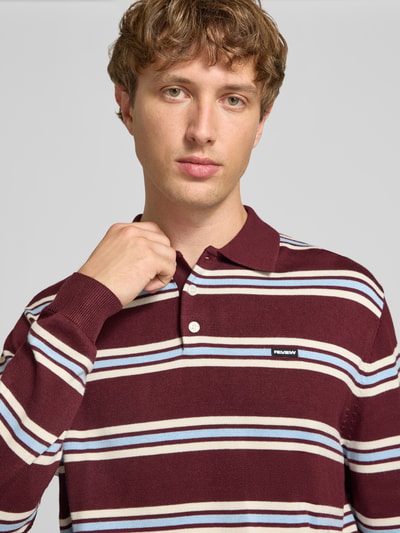 Männermodel trägt Strickpullover mit Polokragen von REVIEW, weinrote Grundfarbe, weiße und blaue Streifen, Label-Stitching, Nahaufnahme Oberkörper.