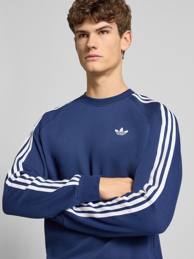 Männermodel trägt Sweatshirt mit Label-Stitching von adidas Originals in Blau mit weißen Streifen, Baumwoll-Mix, Nahaufnahme Oberkörper.