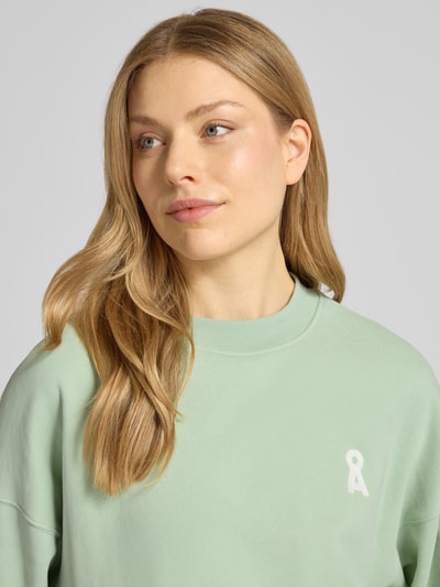 Modelka kobieca nosi Oversized Fit Sweatshirt aus reiner Bio-Baumwolle Modell 'ICONIC A ALIZAA' od Armedangels, pastelowozielony, gładka faktura, logo-stitching, okrągły dekolt, długi rękaw, wykonany z czystej bio-bawełny; zbliżenie na górną część ciała.