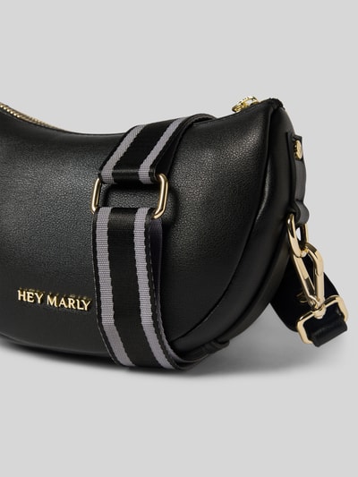 Produktfoto, schwarze Handtasche mit Label-Applikation Modell 'Sporty Sister' von Hey Marly, Textur leicht genarbt, goldfarbene Details, Hauptfach mit Reißverschluss, Schulterriemen aus Stoff, Breite 21 cm, Länge 14 cm, Tiefe 7 cm, Länge Schulterriemen 118 cm.