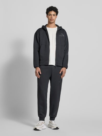 Männermodel trägt schwarze Sweatjacke mit Kapuze von adidas Sportswear, Material-Mix mit Baumwoll-Anteil, Ganzkörperansicht von vorne.