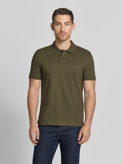 Męski model nosi Slim Fit Poloshirt aus Baumwoll-Mix Modell 'PASSENGER' od BOSS Orange w kolorze oliwkowym, krój slim fit, logo-patch, polokragen, 2-knopf-vorrschlus, krótki rękaw, bawełniany miks, unikalna faktura tkaniny, Ganzkörperansicht von vorne.