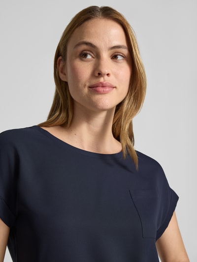 Frauenmodel trägt Blusenshirt mit verlängertem Rücken und Brusttasche von monari, dunkelblauer Stoff, Rundhalsausschnitt, Aufschlag an kurzen Ärmeln, Brusttasche, Label-Detail, Gerader-Schnitt, Damen, Nahaufnahme Oberkörper.