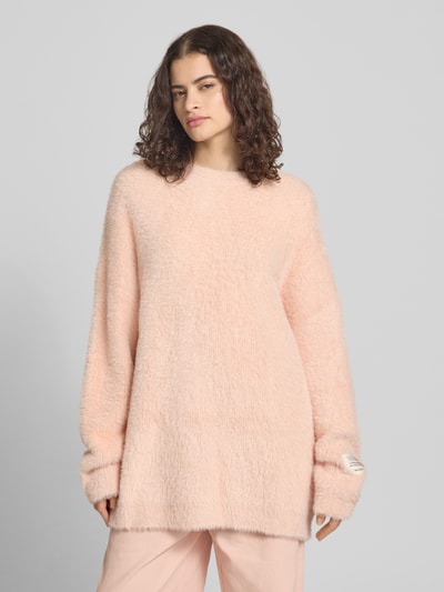 Frauenmodel trägt Oversized Strickpullover mit Label-Detail von Karo Kauer in hellem Rosa, grobe Textur, Rundhalsausschnitt, überschnittene Schultern, Ganzkörperansicht von vorne.