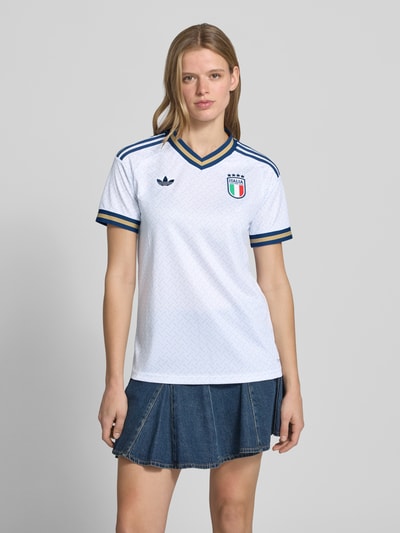Frauenmodel trägt Italien Auswärtstrikot 2026 von adidas Sportswear, weißes mehrfarbiges Kurzarmtrikot mit V-Ausschnitt, Logo-Stitching, Logo-Patch und Kontraststreifen, Regular Fit, Ganzkörperansicht von vorne.