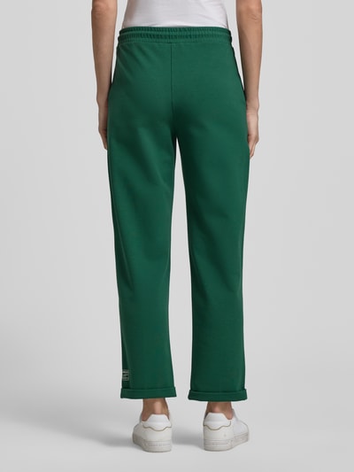 Frauenmodel trägt grüne Straight Fit Stoffhose mit Ziernähten von Marc Cain aus Baumwoll-Modal-Mix, elastischer Bund, fixierte Beinabschlüsse, 7/8-Länge, Ganzkörperansicht Rückansicht.