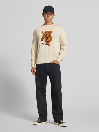 Männermodel trägt Relaxed Fit Strickpullover aus reiner Baumwolle von REVIEW in Beige mit Bärenmotiv, gerippte Abschlüsse, Ganzkörperansicht von vorne.