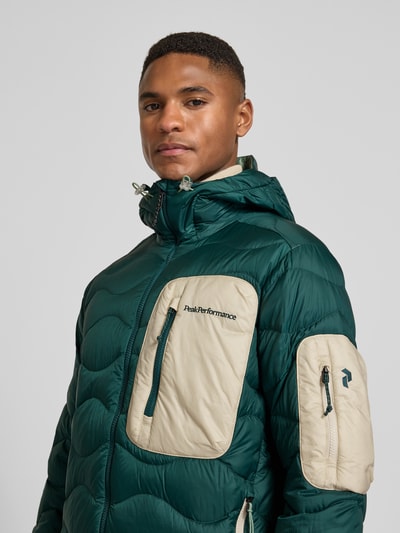 Männermodel trägt Daunenjacke mit Kapuze von Peak Performance in Dunkelgrün mit beigen Reißverschlusstaschen, Nahaufnahme Oberkörper. Gerader Schnitt, Tunnelzug, Brusttasche und seitliche Taschen mit Reißverschluss, Maße laut Kontext für Größe M.