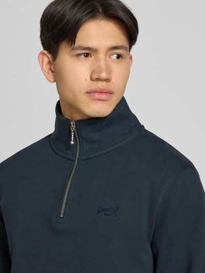 Männermodel trägt Sweatshirt mit Reißverschluss und Label-Stitching von Superdry, mieszanka bawełny, krój regular fit, stójka, wyhaftowane logo, długość tyłu w rozmiarze M 67 cm, długość rękawów w rozmiarze M 69 cm, ciemny kolor, gładka tekstura, nahaufnahme górnej części ciała.
