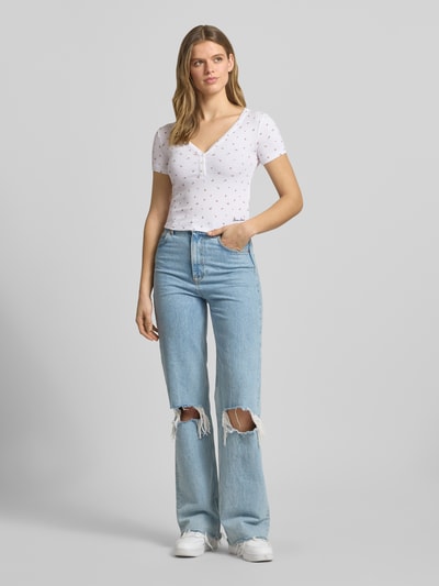Vrouwenmodel draagt Cropped T-shirt met V-hals en knoopsluiting van Guess Jeans, materiaalmix met viscose, kort, logoprint, ruglengte maat S 43 cm, lichte kleuren, gladde textuur, geheel van voren.