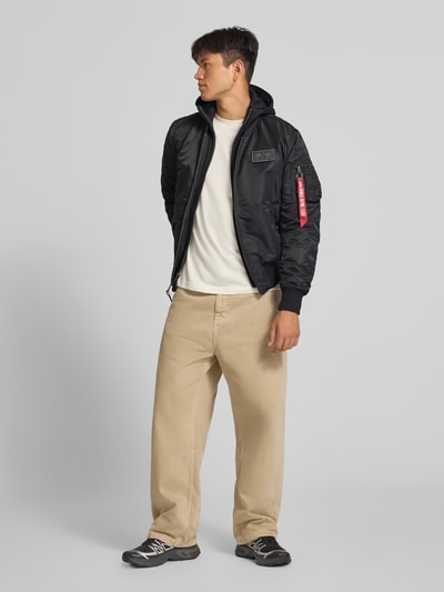 Männermodel trägt schwarze Jacke mit Reißverschlusstasche am Ärmel von Alpha Industries, Regular Fit, Ganzkörperansicht von vorne.