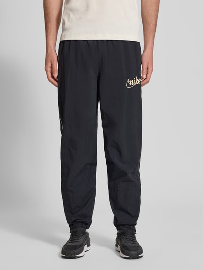 Männermodel trägt Trainingshose mit Logo und elastischem Bund von Nike, Relaxed-Fit, schwarze Farbe, Beinabschlüsse: elastisch, Ganzkörperansicht von vorne. Innenbeinlänge bei Größe M: 76 cm.