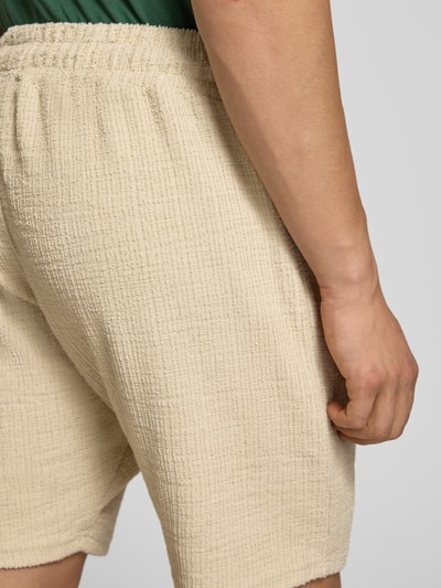 Mężczyzna nosi Shorts mit Label-Patch Modell 'LIBCO' od Pegador, beżowy materiał-mix ze strukturą, elastyczny pas, detailaufnahme biodra. Szerokość w rozmiarze M: 38 cm, długość wewnętrzna nogawki: 23 cm.