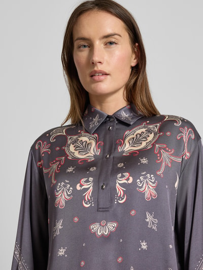 Modelka nosi Blusenshirt mit kurzer Knopfleiste von Marc Cain z czystej wiskozy, luźny krój, kołnierzyk koszulowy, krótka listwa guzikowa, wzory w odcieniach szarości i czerwieni. Zbliżenie na górną część ciała.