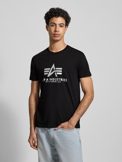 Męski model nosi czarne T-Shirt mit Label-Print von Alpha Industries z nadrukiem, prosty krój, okrągły dekolt, przód w kadrze, bawełna.