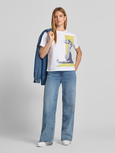 Frauenmodel trägt T-Shirt mit Motiv-Print und Rundhalsausschnitt von Marc Cain aus reiner Baumwolle im Regular Fit, weiße Grundfarbe mit buntem Print, kombiniert mit blauer Jeans und weißem Schuh; Ganzkörperansicht von vorne. Rückenlänge bei Größe 36: 58 cm.