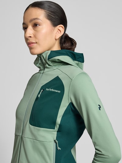 Frauenmodel trägt Jacke mit Logo-Stitching von PEAK PERFORMANCE in Grün mit Elastischer-Material-Mix, Kapuze, Reißverschluss, Brusttasche, körpernahe Passform, Nahaufnahme Oberkörper.
