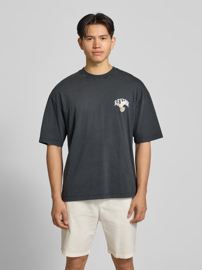 Männermodel trägt T-Shirt mit Logo- und Motiv-Print von REVIEW, schwarze Reine Baumwolle, Loose-Fit, gerippter Rundhalsausschnitt, Ganzkörperansicht von vorne.