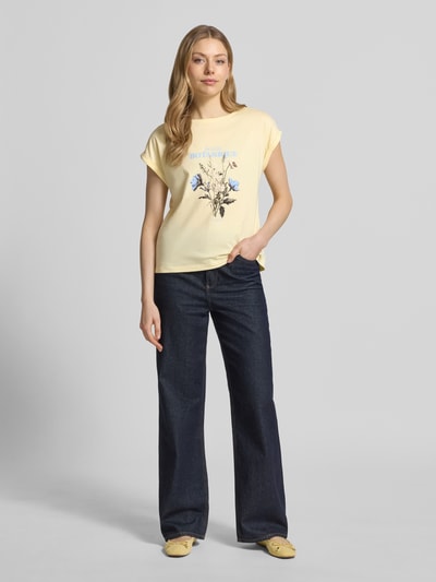 Vrouwenmodel draagt Relaxed Fit T-shirt uit puur katoen van s.Oliver RED LABEL in effen kleuren met print aan de voorkant, korte mouw, ronde hals, labelprint, details: omslag aan de mouw, relaxed fit pasvorm, textuur: zacht katoen, kleur: lichtgeel, voorkant geheel in beeld, Ganzkörperansicht von vorne.
