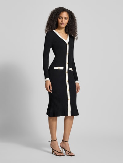 Vrouwenmodel draagt Slim Fit Tricotjurk uit viscosemix model 'Laurel' van Guess in zwart met witte details, doorlopende knoopsluiting, opgestikte zakken, V-hals, lang mouwdesign, ruglengte maat S 110 cm, borstwijdte 33 cm, mouwlengte 63 cm, geheel vooraanzicht.