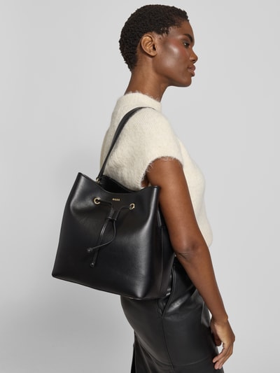 Frauenmodel trägt Henkeltasche mit Label-Applikation Modell 'SANDY BUCKET' von BOSS in Optik: Unifarben, glatte Kunstleder-Textur, Tragehenkel, rechteckige Form, Details wie Reißverschluss, Tunnelzug, Aufteilung: Hauptfach und Innenfach, Standfüße, Nahaufnahme Oberkörper.