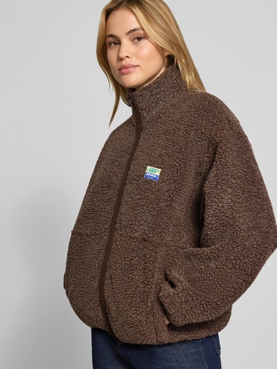 Frauenmodel trägt Regular Fit Jacke aus Teddyfell Modell 'Hoktown' von American Vintage in Braun mit Stehkragen, Logo-Patch, Eingrifftaschen, Reißverschluss, weiche strukturierte Oberfläche, Ganzkörperansicht von vorne. Rückenlänge 62 cm, Schulterbreite 54 cm, Brustweite 46 cm, Ärmellänge 55 cm bei XS/S.