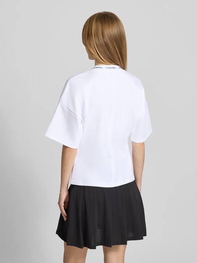Frauenmodel trägt weißes Comfort Fit T-Shirt mit Label-Schriftzug von Calvin Klein Jeans, kombiniert mit schwarzem Rock, Ganzkörperansicht Rückansicht. Damen T-Shirt von Calvin Klein Jeans.