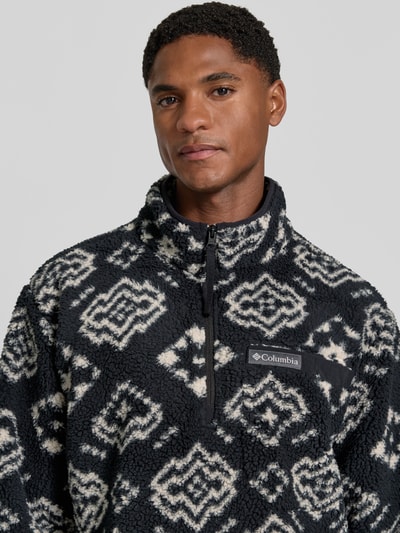 Męski model nosi Regular Fit Pullover mit Logo-Patch od Columbia, Polar, krój regular fit, naszywka z logo, kołnierz zapinany, Długość tyłu w rozmiarze M: 70 cm, Długość rękawów w rozmiarze M: 68 cm, czarno-białe wzory, tekstura polarowa, zbliżenie na górną część ciała.