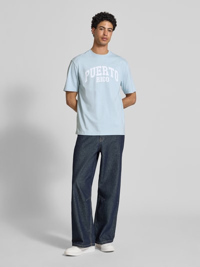 Männermodel trägt Relaxed Fit T-Shirt aus reiner Baumwolle Modell 'RICO' von Only & Sons, hellblau, weißer Print, unifarben, Ganzkörperansicht von vorne.