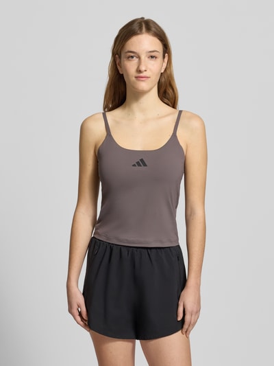 Modelka kobieca ma na sobie Tanktop mit Label-Print od adidas Sportswear, materiał mix z domieszką stretchu, krój slim fit, nadruk z logo, długość tyłu w rozmiarze S: 26 cm, kolor brązowy z czarnym nadrukiem, całkowity widok z przodu.
