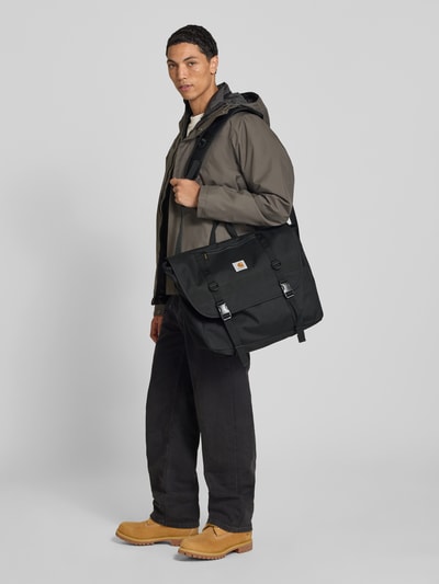 Männermodel trägt Messenger Bag mit Label-Patch Modell 'Jake' von Carhartt Work In Progress in Schwarz mit Klickverschluss, Ganzkörperansicht von vorne.