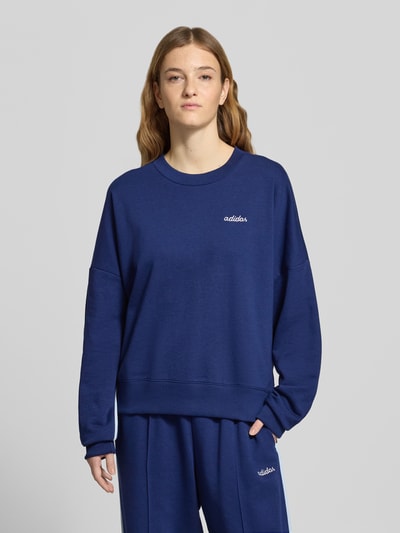 Modelka kobieca nosi Sweatshirt mit Label-Stitching od adidas Sportswear, bawełniany miks z domieszką rayonu, luźny krój, ściągacze, dekolt okrągły, niebieski kolor, haftowane logo, widok całej sylwetki z przodu.