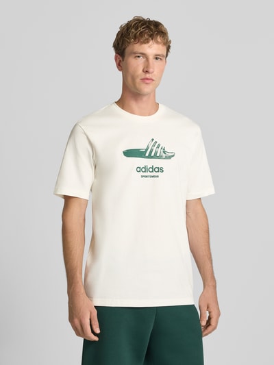 Męski model nosi Regular Fit T-Shirt aus reiner Baumwolle Modell 'SLIDE' von adidas Sportswear w kolorze ecru z zielonym nadrukiem, Ganzkörperansicht von vorne.