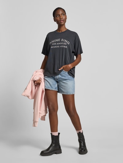 Frauenmodel trägt Relaxed Fit T-Shirt mit Logo-Print von Anine Bing aus reiner Baumwolle, Dunkelgrau, Rundhalsausschnitt, Kurzärmelig, Rückenlänge bei Größe S 59 cm, kombiniert mit Jeansshorts und Stiefeletten, Ganzkörperansicht von vorne.
