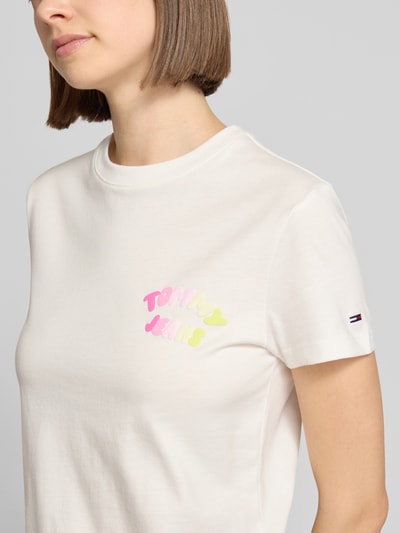 Modelka nosi Regular Fit T-Shirt aus reiner Baumwolle Modell 'BUBBLE' von Tommy Jeans, biała koszulka z nadrukiem label, okrągły dekolt, krótki rękaw, drobny prążkowany materiał, widok z przodu, zbliżenie na górną część ciała.