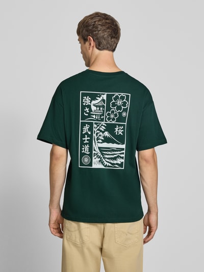 Männermodel trägt Relaxed Fit T-Shirt aus reiner Baumwolle Modell "DYLAN" von JACK & JONES, dunkelgrün mit weißem Print-Detail, Ganzkörperansicht Rückansicht. Rückenlänge bei Größe M: 70 cm, Rundhalsausschnitt.