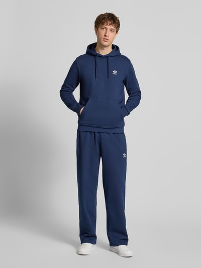 Męski model nosi Hoodie mit Kapuze od adidas Originals, bawełniana mieszanka, krój luźny, kolor granatowy, detal logo, kangurkowa kieszeń, prążkowane wykończenia, długość tyłu w rozmiarze M: 68 cm, długość rękawa w rozmiarze M: 69 cm, widok całej sylwetki z przodu.