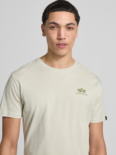 Männermodel trägt T-Shirt aus Baumwolle von Alpha Industries in Beige mit Rundhalsausschnitt und Label-Details, Nahaufnahme Oberkörper.