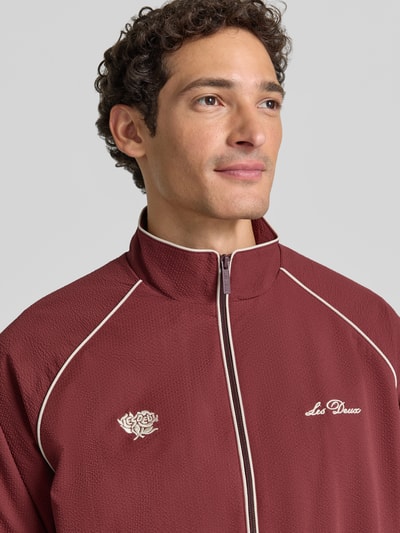 Männermodel trägt bordeauxrote Trainingsjacke mit Label-Stitching Modell 'Seersucker' von Les Deux, Nahaufnahme Oberkörper. Gerader Schnitt, Stehkragen, Kontrastpaspeln, Zwei-Wege-Reißverschluss, elastische Abschlüsse, Rückenlänge bei Größe M: 68 cm, Ärmellänge bei Größe M: 83 cm.